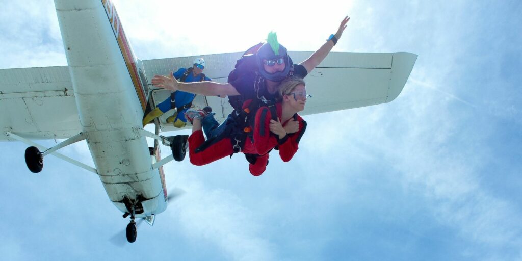 Two people tandem skydiving from a small airplane with a third person jumping behind them. They are in free fall against a bright sky, wearing harnesses and helmets. The airplane is white with landing gear visible.