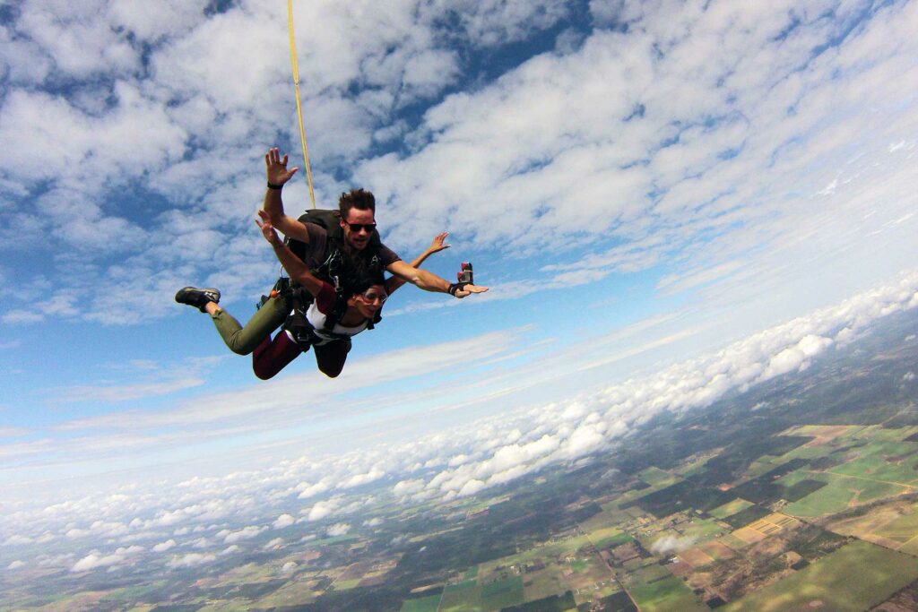 Two people tandem skydiving under a partly cloudy sky, with one holding a camera. They are above a landscape of fields and forests, with the horizon visible in the distance.